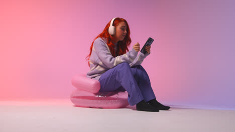 Studio-Shot-Of-Young-Gen-Z-Woman-Wearing-Headphones-Streaming-Music-To-Mobile-Phone